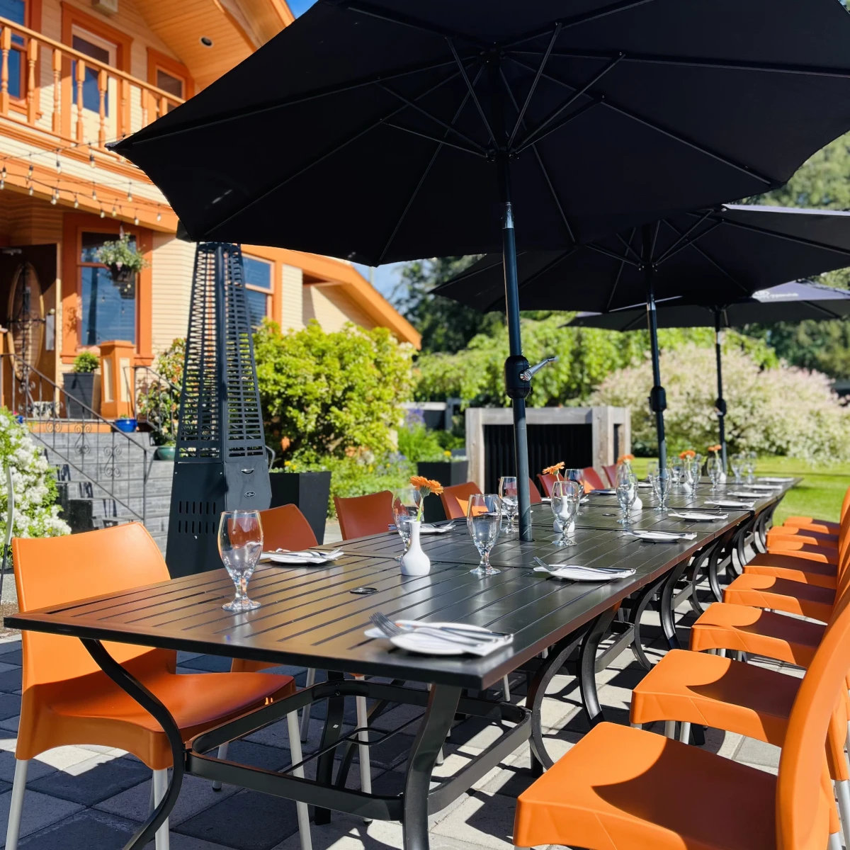 Outdoor dining area with multiple orange and black chairs arranged around square tables. Black umbrellas with logos provide shade. The ground is patterned with alternating grey and charcoal stone tiles. Lush green plants and pink flowers add a pop of color to the scene.