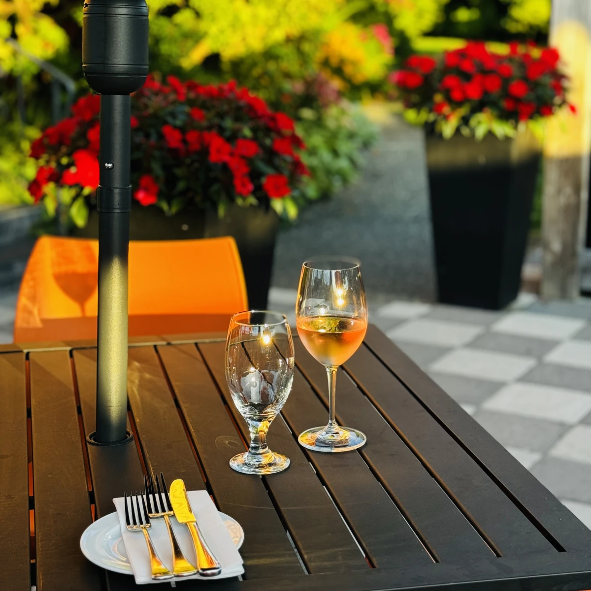 An outdoor dining table set with a bowl of mussels and slices of bread, a glass of rose wine, and an empty wine glass. In the background, is the Mahle House Restaurant with orange siding, surrounded by lush greenery under a clear blue sky.