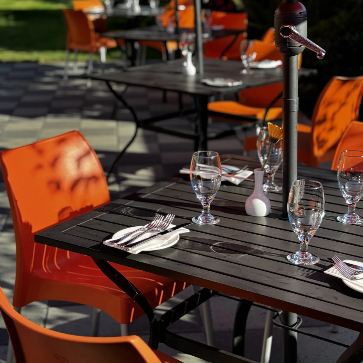 An outdoor patio area with a black umbrella open over a table set with four orange chairs. The patio is bordered by lush green bushes and plants, some with purple and pink flowers. The ground is a mix of grass and grey stone tiles, and there’s a black fence partially visible in the background