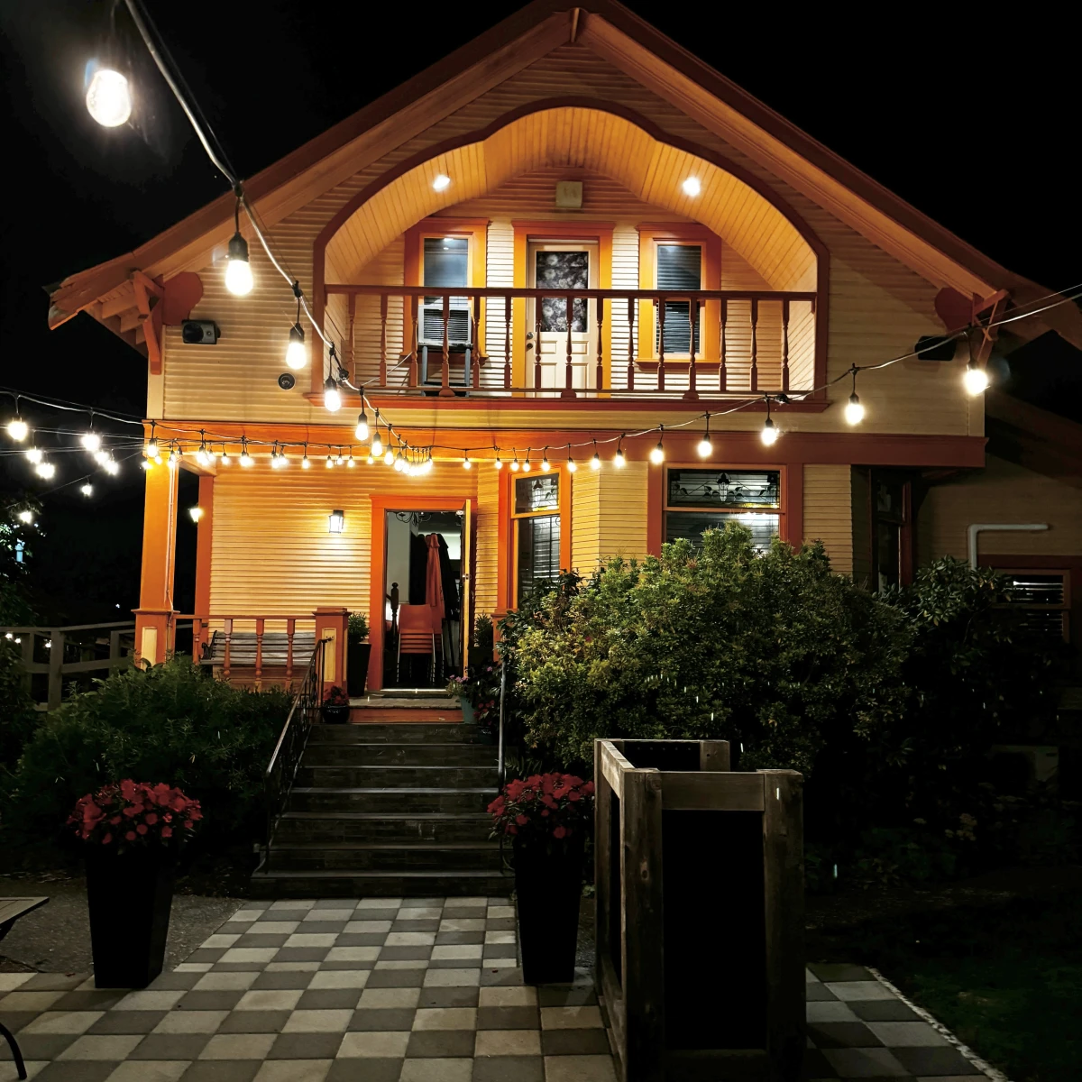 A two-story orange house with a balcony and a front porch adorned with string lights. The house a front door with oval glass, and there is a staircase leading up to the entrance flanked by railings. The front yard features geometric patterned pavement, green shrubbery, and flowers, with a wooden planter box near the sidewalk.