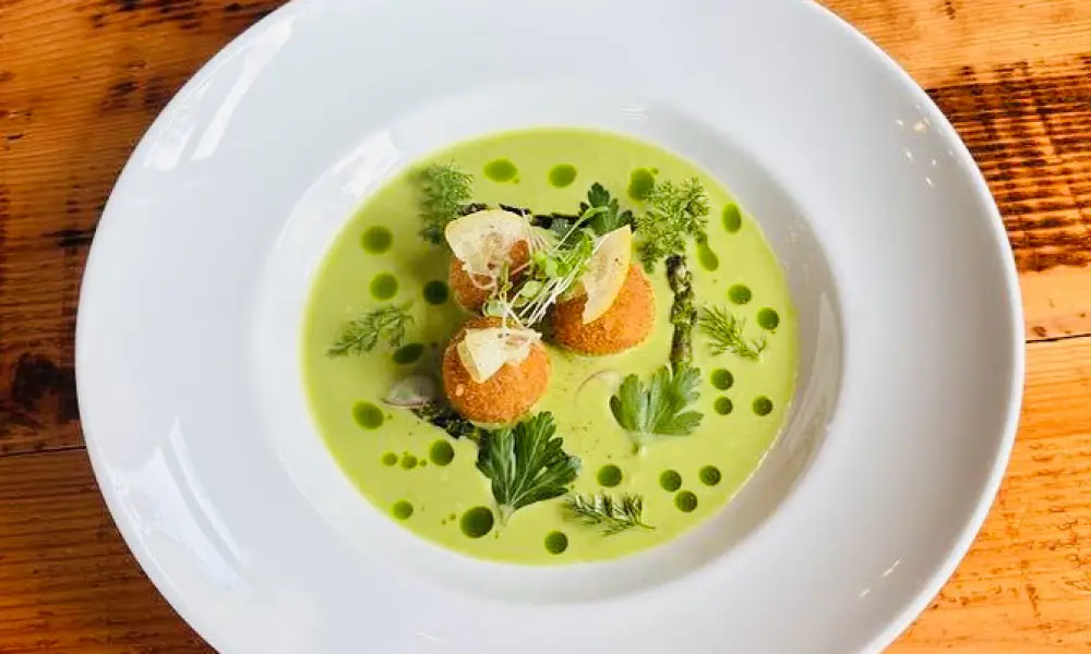 Two round, golden-brown crab cakes topped with a colorful mix of diced vegetables and sprouts, served on a dark slate plate with dollops of creamy sauce and a decorative swipe.