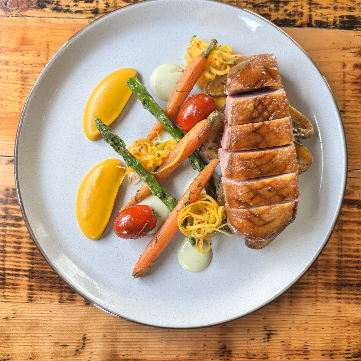 A well-presented gourmet dish on a gray plate featuring a medium-rare cooked steak topped with red cabbage, accompanied by two seared scallops, green beans, asparagus spears, carrot slices, cubed potatoes, and a piece of polenta.