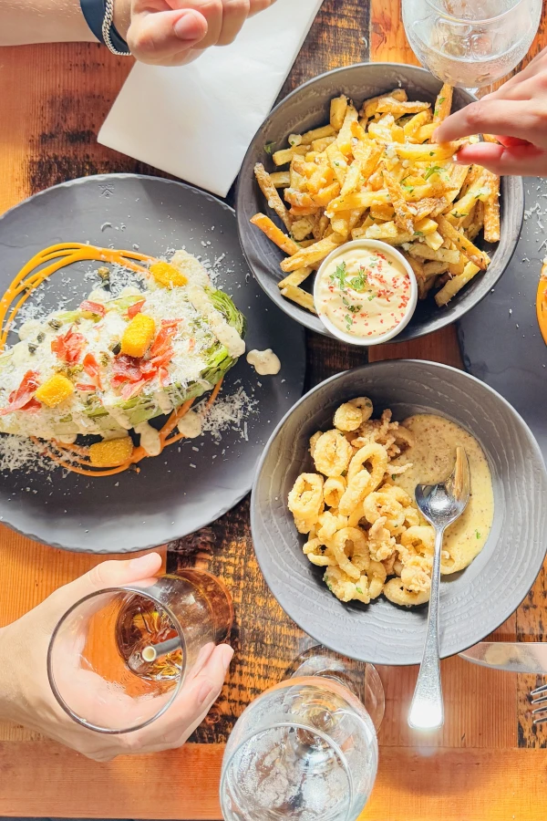 A close-up image of two plates of gourmet food on a kitchen counter, with a chef in the background. The foreground plate is in focus, showcasing a dish with chicken breast topped with herbs, surrounded by an assortment of colorful vegetables including tomatoes and blackberries, and garnished with a yellow sauce