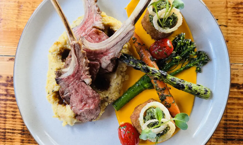 A gourmet dish on a circular dark plate with blue concentric circle patterns. The dish features a golden-brown crusted item, possibly a croquette or fried delicacy, topped with a dollop of cream, orange fish roe, and garnished with purple cabbage and delicate green herbs.