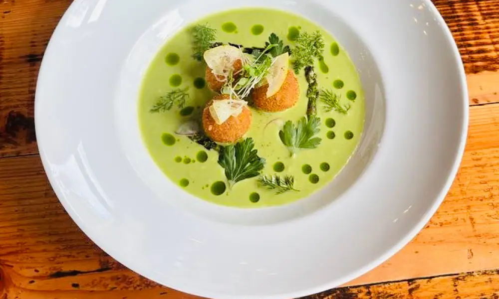 A bowl of steamed clams topped with fresh herbs, slices of cherry tomatoes, and grilled bread. The clams are open and the dish is garnished with cilantro. The bowl sits on a wooden table which adds a rustic feel to the presentation.