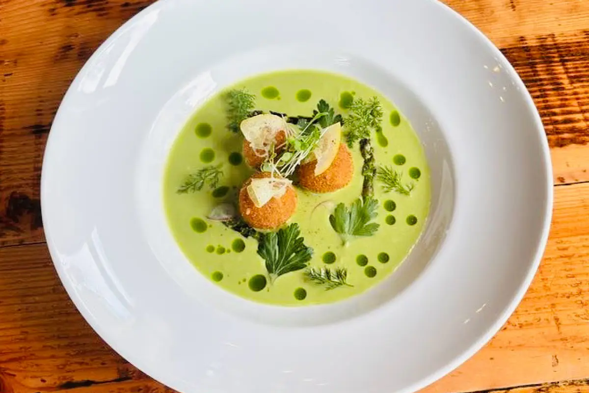 A close-up a dish containing clams in a savory broth, garnished with chopped herbs. In the foreground, there is a piece of grilled bread resting on the rim of the bowl. The background features additional plates with similar content, slightly out of focus, emphasizing the dish in the front.