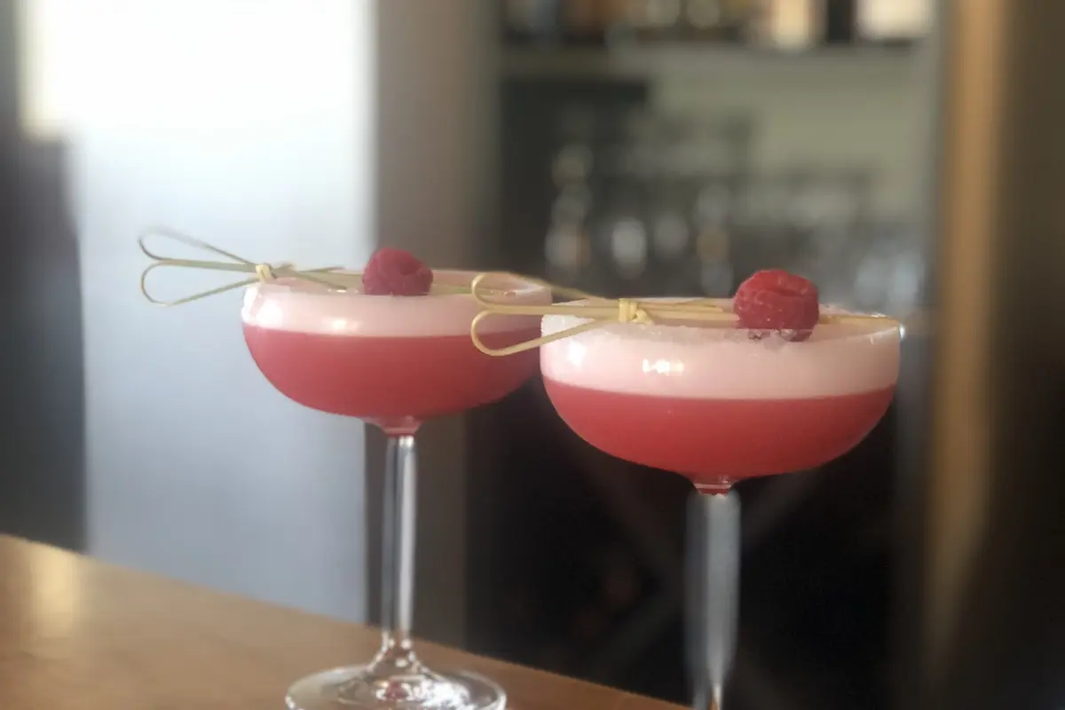 Two pink cocktails in stemmed glasses are presented on a wooden bar counter. Each glass is garnished with a skewered raspberry and a twist of lemon peel resting on the rim. The background features a blurred bar setting with bottles and dim lighting.
